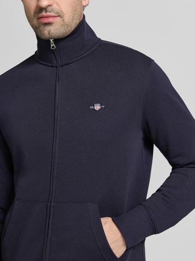 Męski model nosi Regular Fit Sweatjacke mit Stehkragen Modell 'Gield' od Gant, bawełniany miks, granatowy kolor, logo, zamek, kieszeń, elastyczne wykończenia, długość tyłu 69 cm, długość rękawa 64 cm, widok z przodu, zbliżenie górnej części ciała.