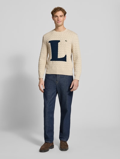 Männermodel trägt Regular Fit Strickpullover aus Baumwoll-Mix von Lacoste in Beige mit großem blauem L, kombiniert mit Jeans, Ganzkörperansicht von vorne. Herren Strickjacke von Lacoste.