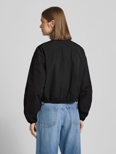 Frauenmodel trägt czarny bluzon 'Relaxed Fit Blouson mit Taillenbändern' von Tommy Jeans, efekt: jednolity kolor, krój relaxed fit, kształt kołnierza: stójka, zapięcie: zamek błyskawiczny, wyhaftowane logo, tasiemka w talii, długi rękaw, zakończenie rękawów: elastyczny, wpuszczane kieszenie z zapięciem na zatrzask; Ganzkörperansicht Rückansicht