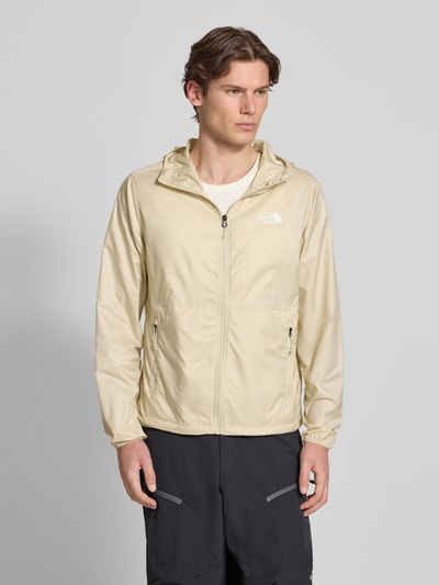 Mannenmodel draagt Windbreaker mit Logo und Reißverschlusstaschen van The North Face in beige met capuchon en ritszakken, regular fit, logoprint, ruglengte bij maat M 66 cm, schouderbreedte 44 cm, borstwijdte 58 cm, mouwlengte 68 cm, voorkant volledige lichaamsweergave.