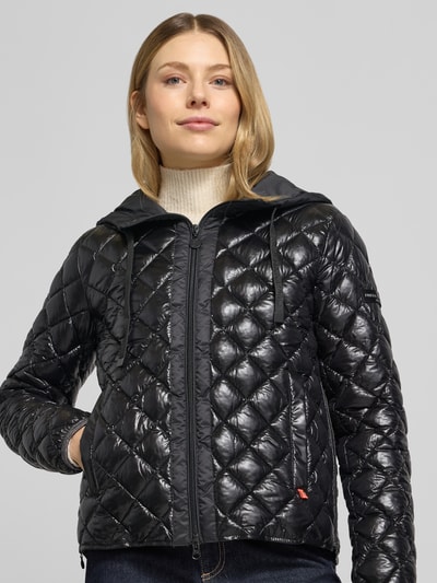 Frauenmodel trägt Regular Fit Jacke in Stepp-Optik Modell 'YOSIE' von Frieda & Freddies, schwarze glänzende Karosteppung, Kapuze mit Tunnelzug, Eingrifftaschen, Reißverschluss, Logo-Applikation, elastische Ärmelabschlüsse, Ganzkörperansicht von vorne.