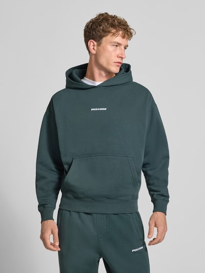 Męski model nosi Oversized Fit Hoodie aus Baumwoll-Mix Modell 'CLARITA' od PEGADOR, kolor zielony, detal z logo, kieszeń kangurka, elastyczne wykończenia, mieszanka bawełny, Ganzkörperansicht von vorne.