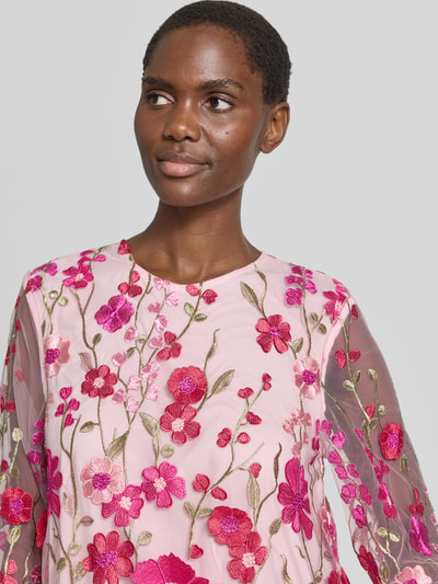 Frauenmodel trägt Knielanges Cocktailkleid mit Stitchings Modell 'FLOWERING' von YAS, semitransparenter Stoff, florale Stickereien in Pink- und Rottönen, Rundhalsausschnitt, Nahaufnahme Oberkörper.