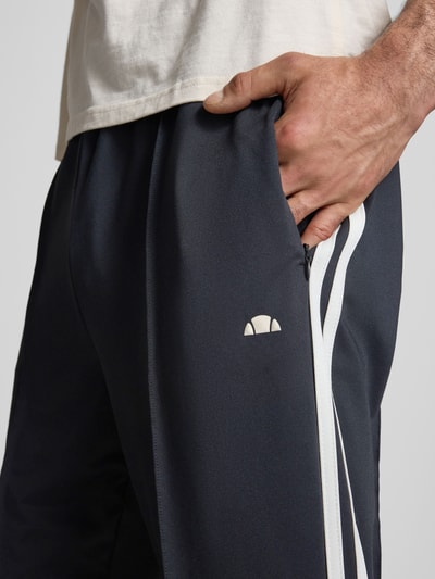 Mężczyzna nosi Straight Fit Sweatpants mit Logo-Detail Modell 'Latucci' od Ellesse, ciemny kolor, białe paski, elastyczny materiał, detal logo, z przodu kieszeń, krój straight fit, zbliżenie na kieszeń, kontekst: męskie spodnie dresowe, długość wewnętrzna 79 cm, elastyczny pas, ozdobne szwy, Ganzkörperansicht von vorne