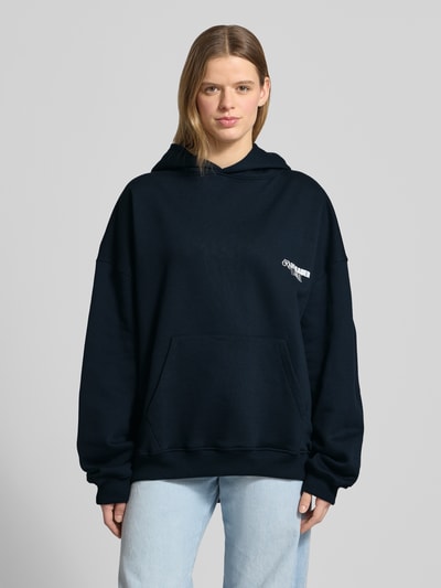 Frauenmodel trägt Oversized Hoodie mit Label-Stitching Modell 'No Hustle' von Karo Kauer, mieszanka bawełny, kaptur, krój oversized fit, kieszeń kangurka, elastyczne wykończenia, wyhaftowane logo, nadruk z napisem z tyłu, kolor ciemnogranatowy, gładka tekstura, Ganzkörperansicht von vorne.