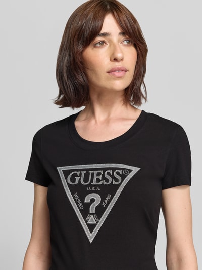 Frauenmodel trägt T-Shirt mit Ziersteinbesatz und Logo von GUESS, schwarz, Baumwoll-Elasthan-Mix, Slim Fit, Nahaufnahme Oberkörper.