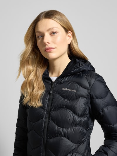 Frauenmodel trägt schwarze Steppjacke mit Daunen-Federn-Füllung Modell 'Helium' von PEAK PERFORMANCE, Nahaufnahme Oberkörper. Regular Fit, Kapuze, Reißverschluss, Label-Stitching, glatte Textur.