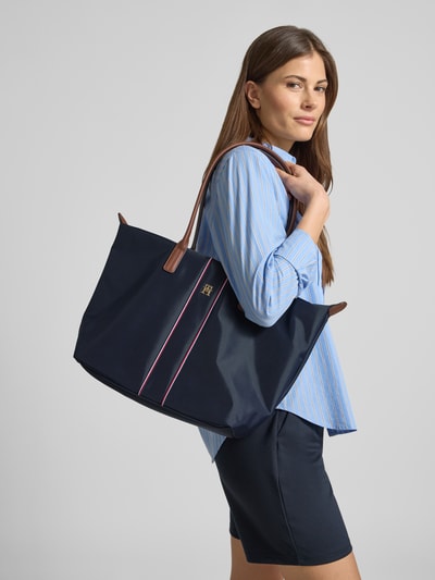 Modelka nosi Henkeltasche mit gestickten Kontraststreifen od Tommy Hilfiger, granatowa, efekt: jednolity kolor, zamek błyskawiczny, główna przegroda, przegródka zapinana na zamek błyskawiczny wewnątrz, aplikacja z logo z metalu, detale: paski w kontrastowym kolorze, widok całej sylwetki z przodu.