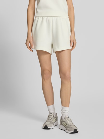 Frauenmodel trägt Shorts mit Label-Print von adidas Sportswear in Offwhite, lockerer Schnitt, Material-Mix, Nahaufnahme Oberkörper.