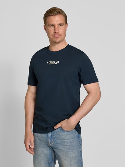 Mężski model nosi Regular Fit T-Shirt aus reiner Baumwolle od s.Oliver RED LABEL, kolor granatowy, krój regular fit, czysta bawełna, krótki rękaw, okrągły dekolt, napis logo z przodu, tkanina dżersej, widok całej sylwetki z przodu.