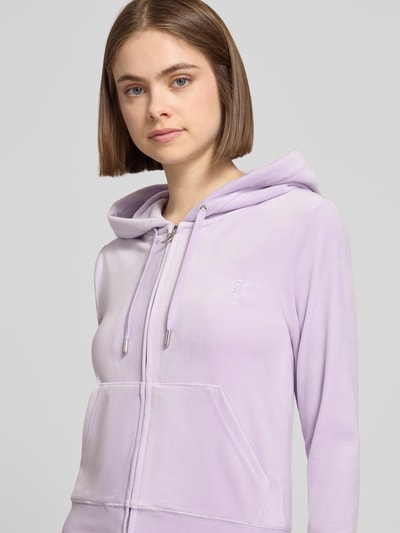 Frauenmodel trägt Slim Fit Sweatjacke aus Samt Modell 'Robertson' von Juicy Couture in Flieder mit Kapuze, Tunnelzug, Reißverschluss, geteilte Känguru-Tasche, elastische Abschlüsse; Elastischer-Material-Mix, Damen Sweatjacke, Nahaufnahme Oberkörper.
