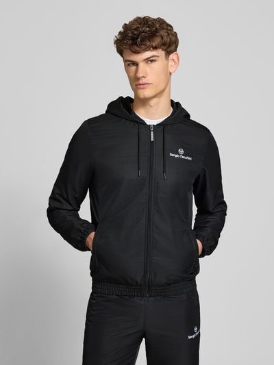Männermodel trägt schwarzen Regular Fit Tracksuit mit Logo-Stitching Modell 'Carson' von SERGIO TACCHINI, Ganzkörperansicht von vorne, Kapuze mit Tunnelzug, Reißverschluss, elastische Abschlüsse, Eingrifftaschen, Bundweite 38 cm, Rückenlänge 67 cm, Schulterbreite 43 cm, Ärmellänge 70 cm, Innenbeinlänge 76 cm.