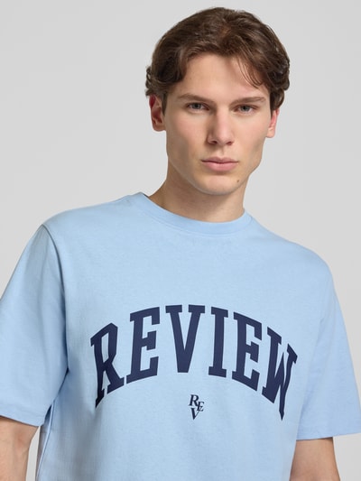 Männermodel trägt T-shirt mit labelprint en ronde hals van REVIEW in lichtblauw, puur katoen, regular fit, ruglengte maat M 69 cm, nahaufnahme oberkörper.