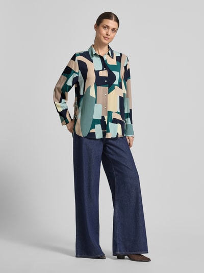 Frauenmodel trägt Relaxed Fit Hemdbluse aus reiner Viskose von Tom Tailor mit abstraktem Allover-Muster, Hemdblusenkragen, Schleifen-Details, Knopfleiste und langen Ärmeln, kombiniert mit weiter Jeans; Ganzkörperansicht von vorne.