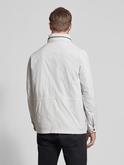 Männermodel trägt Regular Fit Jacke mit stójka od Bugatti w kolorze jasnoszarym, aplikacja z logo, długość tyłu 72 cm, szerokość ramion 46 cm, szerokość w rozmiarze 50: 52 cm, długość rękawów 68 cm, kieszenie na piersi, schowany kaptur, tkanina o gładkiej fakturze, całkowity widok z tyłu.