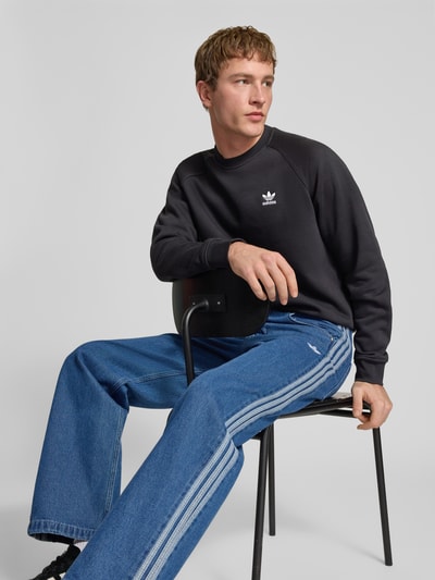 Męski model nosi Regular Fit Sweatshirt aus Baumwoll-Mix od adidas Originals, bawełniany czarny sweter z logo, okrągły dekolt, prążkowane wykończenia, gładka struktura, krój regular fit, zdjęcie całej sylwetki z przodu.