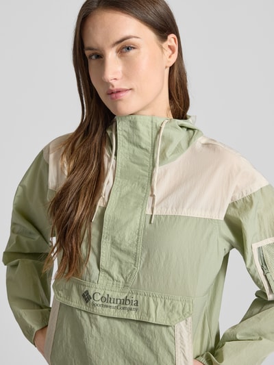 Frauenmodel trägt Regular Fit Windbreaker mit Logo-Print Modell 'Challenger' von Columbia in hellem Grün-Beige, Struktur aus glattem Stoff, mit Kapuze und Känguru-Tasche, Omni-Shield™, Nahaufnahme Oberkörper.