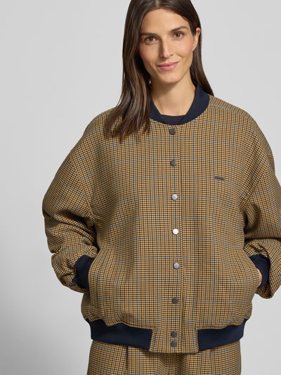 Frauenmodel trägt Loose Fit Bomberjacke mit Knopfleiste Modell 'chicca' von Gestuz in kariertem Beige-Braun mit Label-Stitching, Ganzkörperansicht von vorne. Damenjacke mit Stehkragen, Eingrifftaschen und elastischen Abschlüssen, Maße laut Kontext.