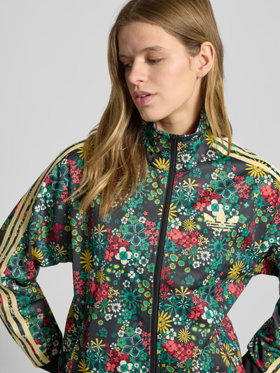 Vrouwenmodel draagt Regular Fit Jacke met Logo-Stitching Modell 'Firebird' van adidas Originals met florale allover-print, staande kraag, steekzakken, elastische boorden, ritssluiting, kleurrijke bloemen in groen, geel en rood, detailopname bovenlichaam.