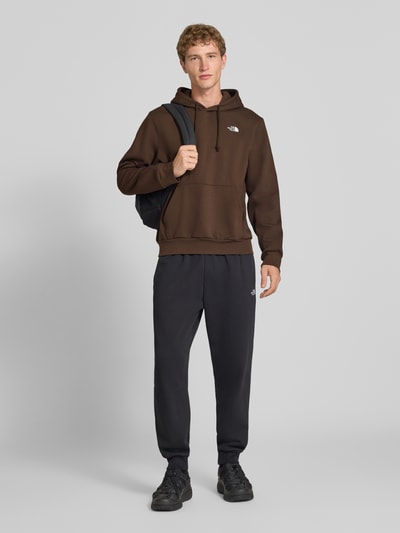 Mannenmodel draagt Regular Fit Hoodie met kangoeroezak Model "EVOLUTION" van The North Face in katoenmix, logostitching, bruin en zwart, ruglengte bij maat M 67 cm, mouwlengte maat M 67 cm, full body vooraanzicht.