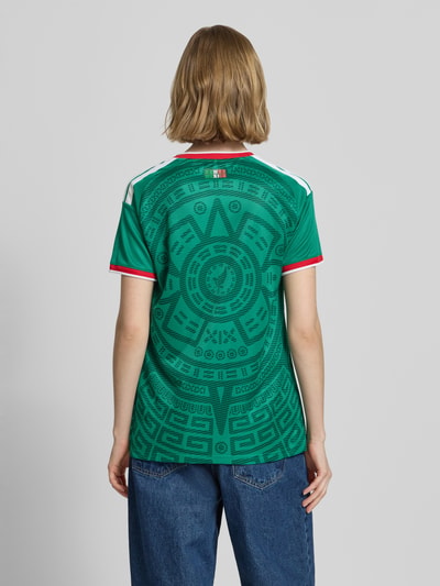Frauenmodel trägt Mexiko Heimtrikot 2026 von adidas Sportswear, Mehrfarbig, Regular-Fit, FMF-Applikation, Kontraststreifen, Logo-Stitching, V-Ausschnitt, Ärmelform: Kurzarm, Ganzkörperansicht Rückansicht.