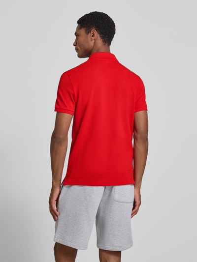 Männermodel trägt rotes Slim Fit Poloshirt aus reiner Baumwolle von Lacoste, kombiniert mit grauer Shorts, Ganzkörperansicht Rückansicht.