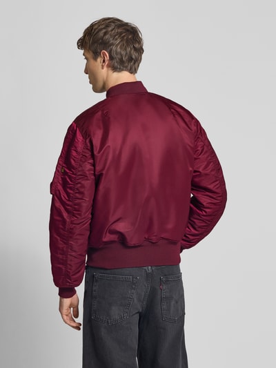 Męski model nosi bomberkę z stójką, krój regular fit, model 'Heritage' od Alpha Industries, bordowy kolor, widok całej sylwetki od tyłu. Długość tyłu w rozmiarze M: 63 cm, szerokość ramion w rozmiarze M: 50 cm, szerokość klatki piersiowej w rozmiarze M: 57 cm, kieszenie z patką, elastyczne wykończenia, zamek błyskawiczny.