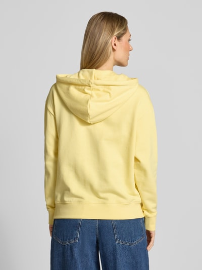 Modelka kobieca nosi Hoodie mit Logo-Stickerei Modell 'LEA' od Mustang z bawełny, luźny krój, żółty kolor, kieszeń kangurka, wyhaftowane logo, kaptur z tunelem, prążkowane wykończenia, widok całej sylwetki od tyłu.