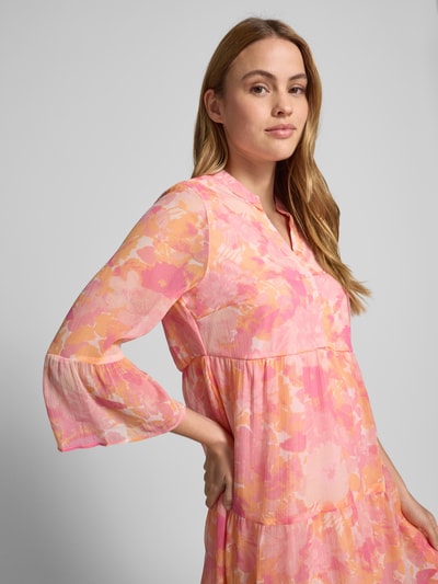 Kobiece modelka nosi sukienkę mini z szyfonu o wzorze na całej powierzchni, model 'Smilla' od Vero Moda, taliowany krój, dekolt w serek, rękawy z falbanami, efekt lekko prześwitującej tkaniny, kolor różowo-pomarańczowy, detaliczne wykończenie materiału, zbliżenie na górną część ciała, przód.