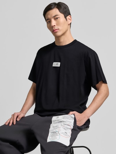 Männermodel trägt Relaxed Fit T-Shirt mit Logo-Detail von MM6 Maison Margiela aus schwarzer Baumwolle, Rundhalsausschnitt, Nahaufnahme Oberkörper.