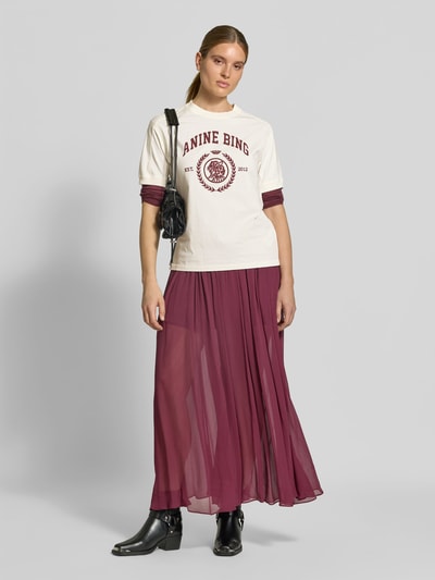Frauenmodel trägt Regular Fit T-Shirt aus reiner Baumwolle von Anine Bing mit Logo-Print, kombiniert mit langem, bordeauxfarbenem Rock, Ganzkörperansicht von vorne.