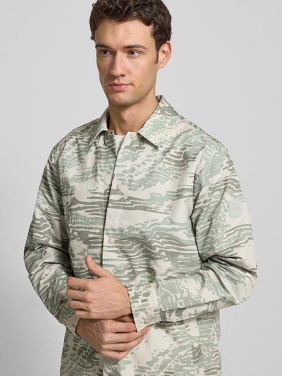 Männermodel trägt Regular Fit Hemdjacke mit Rumpftunnelzug Modell 'LELDON' von BOSS Orange, Allover-Camouflage-Muster, Nahaufnahme Oberkörper.