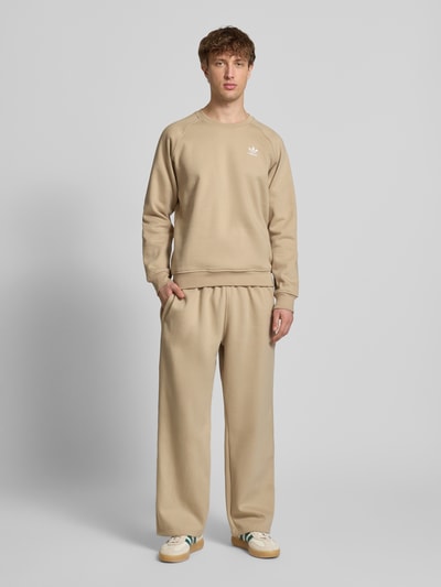 Mężczyzna nosi Wide Fit Sweatpants mit elastischem Bund od adidas Originals, kolor beżowy, bawełniany miks, logo, szerokie nogawki, Ganzkörperansicht von vorne.