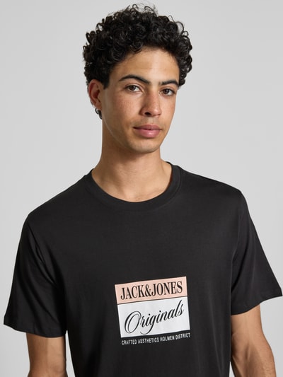 Męski model nosi męski t-shirt z nadrukiem z napisem z czarnej woskowanej bawełny, model 'RHYERES' od Jack & Jones, krój regular fit, okrągły dekolt, długość tyłu w rozmiarze M: 67 cm, zbliżenie górnej części ciała.
