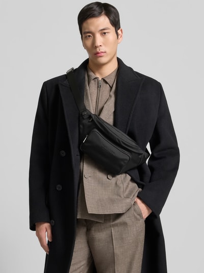 Männermodel trägt Bauchtasche mit Label-Detail von Dsquared2 in Schwarz, dezente Struktur, Reißverschlüsse und Riemen sichtbar, Ganzkörperansicht von vorne. Hauptfach, Nebenfach und Maße laut Kontext.