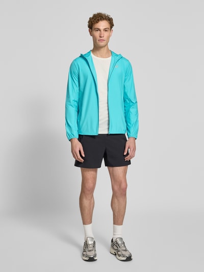 Männermodel trägt Windbreaker mit Logo-Print und Kapuze von ASICS in Türkis, gerader Schnitt, Reißverschluss, Ganzkörperansicht von vorne.