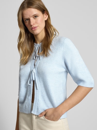 Modelka kobieca ma na sobie Regular Fit Cardigan mit Schnürungen Modell 'SIMONI' od Only, jasnoniebieski, gładka dzianina, krój regular fit, półrękaw, okrągły dekolt, detale ze sznurkami i ściągaczem, widok z przodu na całą sylwetkę.