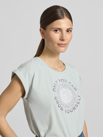 Modelka kobieca nosi damski t-shirt z nadrukiem z napisem od G.I.G.A. DX, mieszanka bawełny, krój regular fit, okrągły dekolt, długość tyłu w rozmiarze 36: 61 cm, jasne kolory, tekstura gładka, zbliżenie na górną część ciała.