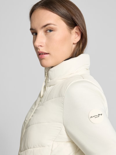 Kobiece modelka nosi Slim Fit Steppjacke mit gefüttertem Stehkragen od Christian Berg Woman Selection, jasny kremowy kolor, pikowana tekstura, wysoki kołnierz, detal logo na ramieniu, krój slim fit, zbliżenie na górną część ciała z profilu. Szerokość w rozmiarze 36: 51 cm, długość pleców: 60 cm, szerokość ramion: 41 cm, długość rękawa: 62 cm.