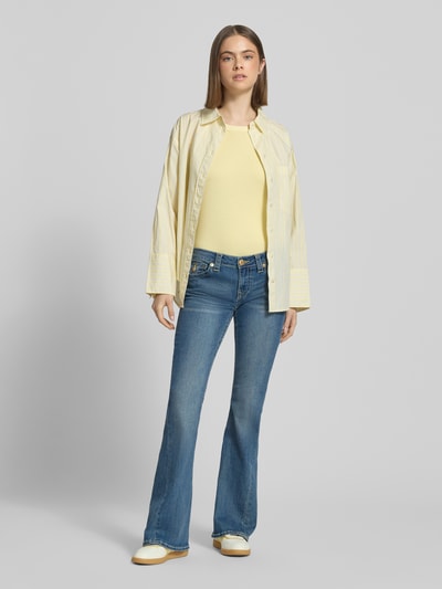 Frauenmodel trägt Low Rise Flared Jeans mit Eingrifftaschen Modell 'JOEY' von TRUE RELIGION im Used-Look aus Baumwoll-Mix mit Stretch-Anteil, mittelblaue Farbe, Flared-Cut, Ganzkörperansicht von vorne.