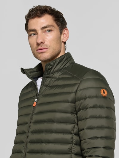 Männermodel trägt olivgrüne Steppjacke mit Zweiwege-Reißverschluss Modell 'ALEXANDER' von Save The Duck, leichte Struktur, Nahaufnahme Oberkörper. Herren Steppjacke im Slim Fit mit Stehkragen, Reißverschlusstaschen, Größe M: Rückenlänge 67 cm, Schulterbreite 47 cm, Brustweite 57 cm, Ärmellänge 68 cm.