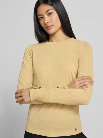 Frauenmodel trägt Regular Fit Longsleeve mit Rundhalsausschnitt von Marc Cain in Beige mit Glitzer-Optik, Ziersteinbesatz, Logo-Detail, elastischer Material-Mix, Ganzkörperansicht von vorne.