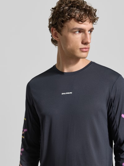 Männermodel trägt Funktionsshirt mit Logo Modell 'Kout' von Salomon in Dunkelgrau mit Rundhalsausschnitt, Nahaufnahme Oberkörper. Regular Fit, Longsleeve, Rückenlänge bei Größe M 70 cm.