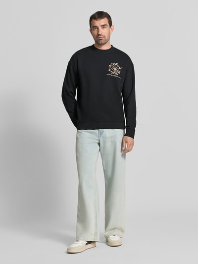 Männermodel trägt Relaxed Fit Pullover mit Logo-Print von Scotch & Soda in zwart, puur katoen, ronde hals, elastische boordjes, ganzkörperansicht von vorne.