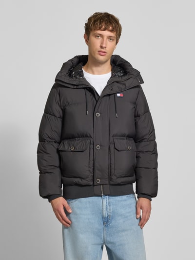 Männermodel trägt Regular Fit Pufferjacke mit Kapuze von Tommy Jeans, unifarben, Logo-Patch, Steppstruktur, Ganzkörperansicht von vorne.