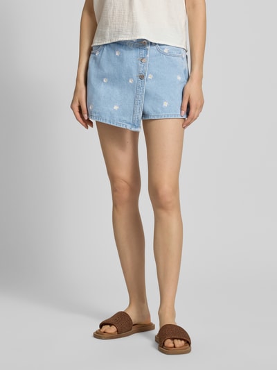 Frauenmodel trägt Regular Fit Skort aus reiner Baumwolle Modell 'LESLY' von Only in hellblauem Denim mit Stickerei, Wickeloptik, seitliche Eingrifftaschen, Gürtelschlaufen, Knopfleiste und verdecktem Reißverschluss, Ganzkörperansicht von vorne.