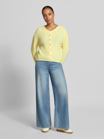 Frauenmodel trägt Relaxed Fit Strickjacke aus Woll-Mix mit Leinen-Anteil von American Vintage in Hellgelb, gerippte Bündchen, Ganzkörperansicht von vorne, Damen Strickjacke, lange Ärmel, durchgehende Knopfleiste, weiche Textur, Rückenlänge 52 cm, Ärmellänge 56 cm.
