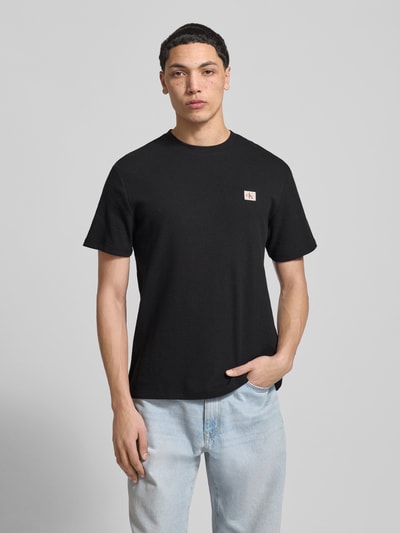 Männermodel trägt Regular Fit T-Shirt mit Logo-Patch von Calvin Klein Jeans, schwarzer Baumwoll-Mix, Strukturmuster, Rundhalsausschnitt, Ganzkörperansicht von vorne.