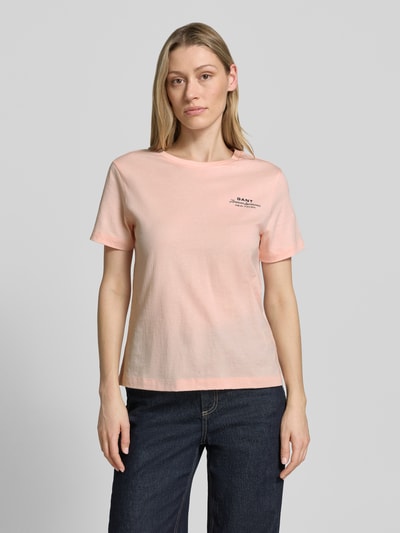 Kobiecy model nosi T-Shirt mit Label-Print und Rundhalsausschnitt od Gant, bawełna, krój regular fit, długość tyłu w rozmiarze S: 57 cm, jasnoróżowy, gładka faktura, nadruk z logo, okrągły dekolt, całkowity widok z przodu.
