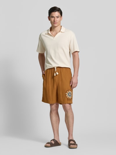 Männermodel trägt Straight Fit Shorts aus Leinen-Viskose-Mix mit Motiv-Stitching von Scotch & Soda, Logo-Stitching, Tunnelzug, Ganzkörperansicht von vorne.